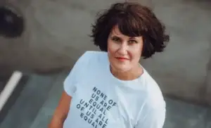 Ein Porträt einer Frau mit dunklen, lockigen Haaren, die direkt in die Kamera blickt. Sie trägt ein weißes T-Shirt mit der Aufschrift: „NONE OF US ARE EQUAL UNTIL ALL OF US ARE EQUAL“ (Keiner von uns ist gleichgestellt, bis wir alle gleichgestellt sind). Die Aufnahme erfolgt aus einer leichten Vogelperspektive vor einem neutralen, grau-bräunlichen Hintergrund. Ihr Gesichtsausdruck ist ruhig und bestimmt.