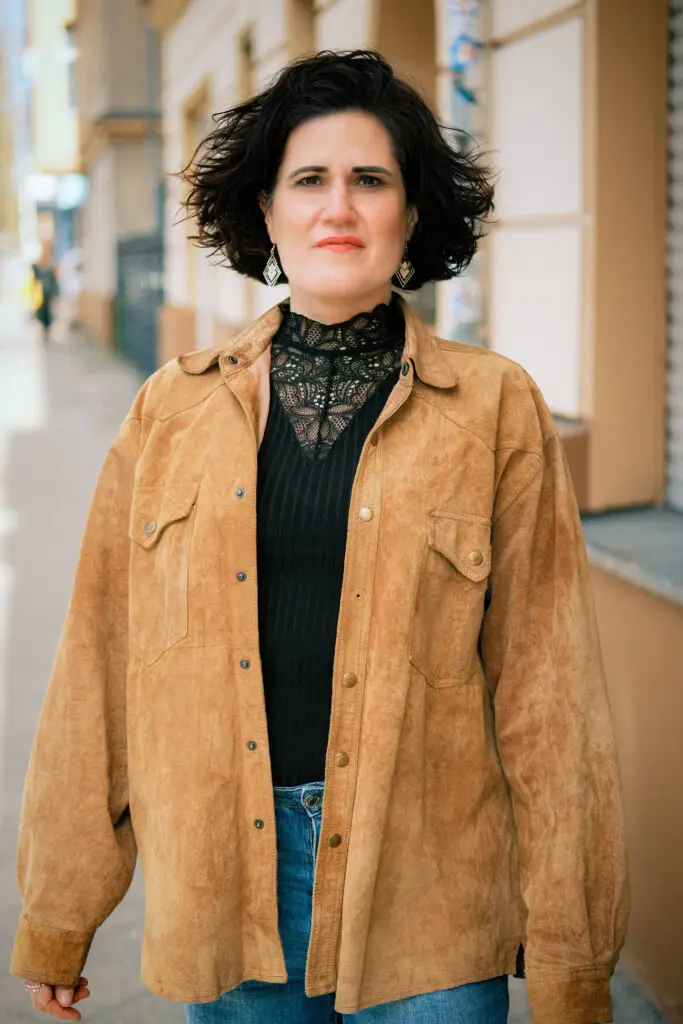 Porträtaufnahme von Stefanie López, die in einer Stadtstraße steht. Sie trägt eine locker sitzende, braune Wildlederjacke über einem schwarzen Oberteil mit einem hohen Kragen aus Spitze und blaue Jeans. Ihr dunkles, gewelltes Haar ist offen gestylt, und sie trägt geometrische Ohrhänger.