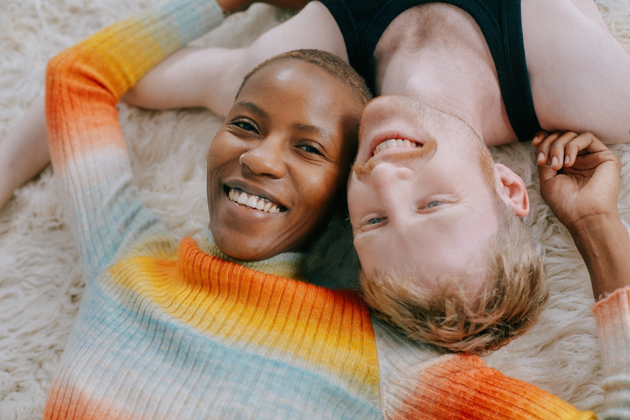 Eine Nahaufnahme von oben: Eine Person mit kurzen Haaren in einem regenbogenfarbenen Strickpullover und ein Mann mit rötlichem Haar liegen Kopf an Kopf auf einem flauschigen weißen Teppich und lachen in die Kamera.