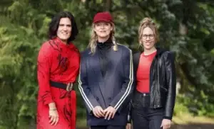 Ein Porträt von drei Frauen, die nebeneinander im Freien vor einem unscharfen, grünen Blätterhintergrund stehen. Die Frau links trägt ein auffälliges, rotes Kleid mit schwarzem Muster und einem breiten schwarzen Gürtel. Die Frau in der Mitte trägt ein dunkelblaues Sakko mit gelben Streifen an den Ärmeln über einem schwarzen Rollkragenpullover, dazu eine rote Basecap und hat ihr blondes Haar zu einem Zopf geflochten. Die Frau rechts trägt eine schwarze Lederjacke über einem roten Oberteil, eine dunkle Hose und eine Brille. Alle drei blicken lächelnd in die Kamera. Das Licht wirkt natürlich und freundlich.