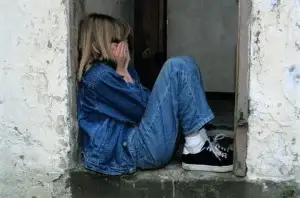 Ein Kind sitzt allein in einer Hauseingangsnische eines baufälligen Gebäudes. Das Kind trägt eine Jeansjacke, Jeans und dunkle Sneaker. Es hat die Beine angezogen und hält sich beide Hände vor das Gesicht, was eine traurige oder verzweifelte Stimmung vermittelt.