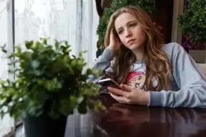 Ein Porträt eines Mädchens mit langen, gewellten Haaren, das an einem dunklen Tisch sitzt. Sie stützt ihren Kopf nachdenklich auf eine Hand und hält in der anderen ein Smartphone. Ihr Blick ist ernst und geht am Betrachter vorbei in die Ferne. Im Vordergrund steht eine unscharfe grüne Pflanze.