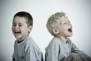 Ein helles Porträt von zwei Kindern, die Rücken an Rücken sitzen und herzhaft lachen. Beide tragen graue Langarmshirts. Das Kind links hat die Augen fest geschlossen und den Mund weit zum Lachen geöffnet, während das Kind rechts den Kopf leicht in den Nacken legt und ebenfalls begeistert lacht.