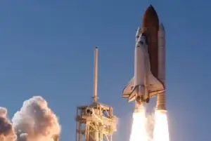Eine beeindruckende Aufnahme eines startenden Space Shuttles. Die Raumfähre hebt mit gleißend hellen Flammen aus den Boostern von der Startrampe ab und steigt in einen klaren, blauen Himmel auf. Links im Bild ist der Startturm zu sehen, der teilweise von einer massiven, weißen Rauch- und Dampfwolke verhüllt wird, die durch die Zündung der Triebwerke entstanden ist. Die Szene vermittelt die enorme Kraft und Dynamik eines Raketenstarts.