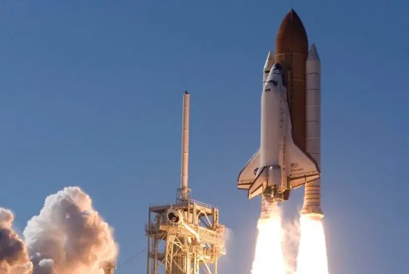 Eine beeindruckende Aufnahme eines startenden Space Shuttles. Die Raumfähre hebt mit gleißend hellen Flammen aus den Boostern von der Startrampe ab und steigt in einen klaren, blauen Himmel auf. Links im Bild ist der Startturm zu sehen, der teilweise von einer massiven, weißen Rauch- und Dampfwolke verhüllt wird, die durch die Zündung der Triebwerke entstanden ist. Die Szene vermittelt die enorme Kraft und Dynamik eines Raketenstarts.