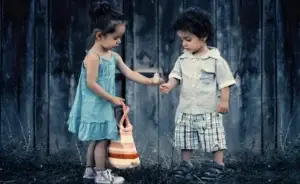 Eine stimmungsvolle Außenaufnahme von zwei kleinen Kindern vor einer dunklen Holzwand. Ein Mädchen in einem hellblauen Kleid und mit einer gestreiften Tasche hält einem Jungen, der ein helles Hemd und karierte Shorts trägt, eine Pusteblume hin. Beide Kinder betrachten die Blume aufmerksam.