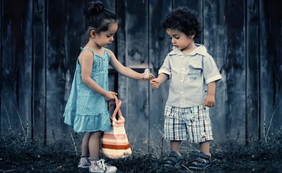 Eine stimmungsvolle Außenaufnahme von zwei kleinen Kindern vor einer dunklen Holzwand. Ein Mädchen in einem hellblauen Kleid und mit einer gestreiften Tasche hält einem Jungen, der ein helles Hemd und karierte Shorts trägt, eine Pusteblume hin. Beide Kinder betrachten die Blume aufmerksam.