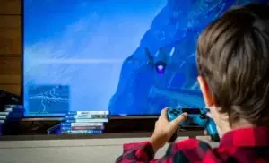 Eine Rückansicht eines Kindes, das auf einem großen Fernseher ein Videospiel spielt. Auf dem Bildschirm ist eine Flugsitzung mit einem Kampfjet in einer bläulichen Gebirgslandschaft zu sehen. Das Kind hält einen blauen Controller in den Händen und trägt ein rot-schwarz kariertes Hemd. Vor dem Fernseher stapeln sich mehrere PS4-Spielehüllen, darunter Titel wie „Far Cry“. Die Szene fängt einen Moment konzentrierter Freizeitbeschäftigung ein.