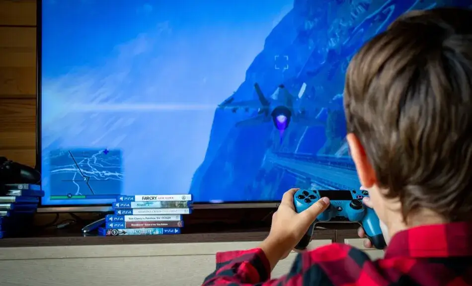 Eine Rückansicht eines Kindes, das auf einem großen Fernseher ein Videospiel spielt. Auf dem Bildschirm ist eine Flugsitzung mit einem Kampfjet in einer bläulichen Gebirgslandschaft zu sehen. Das Kind hält einen blauen Controller in den Händen und trägt ein rot-schwarz kariertes Hemd. Vor dem Fernseher stapeln sich mehrere PS4-Spielehüllen, darunter Titel wie „Far Cry“. Die Szene fängt einen Moment konzentrierter Freizeitbeschäftigung ein.