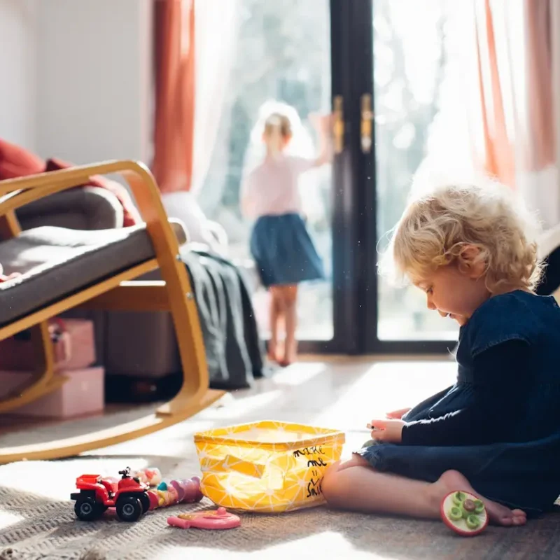 Kleines Kind im Vordergrund kniet auf einem Teppich und spielt mit Spielzeug. Im Hintergrund unscharf ein älteres Kind was aus dem Bodentiefen Fenster guckt.